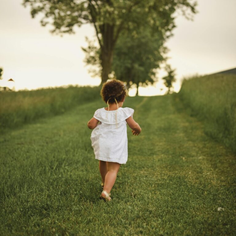 A young child runs freely down a grassy path surrounded by lush trees in a park during summer.