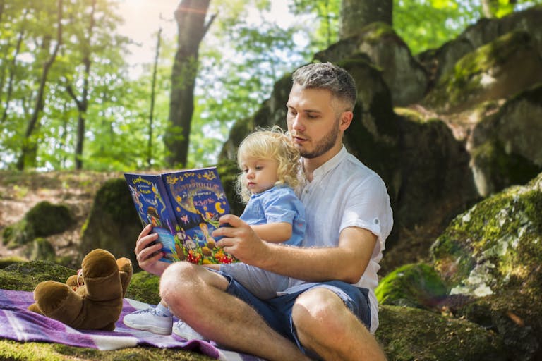 Father and toddler bonding over a book in a sunny forest setting.