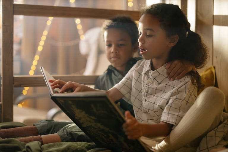 Two siblings enjoying a storybook together, surrounded by cozy lighting indoors.