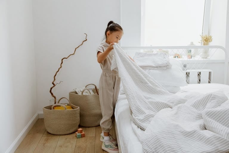 A young girl making her bed in a bright, minimalist bedroom.