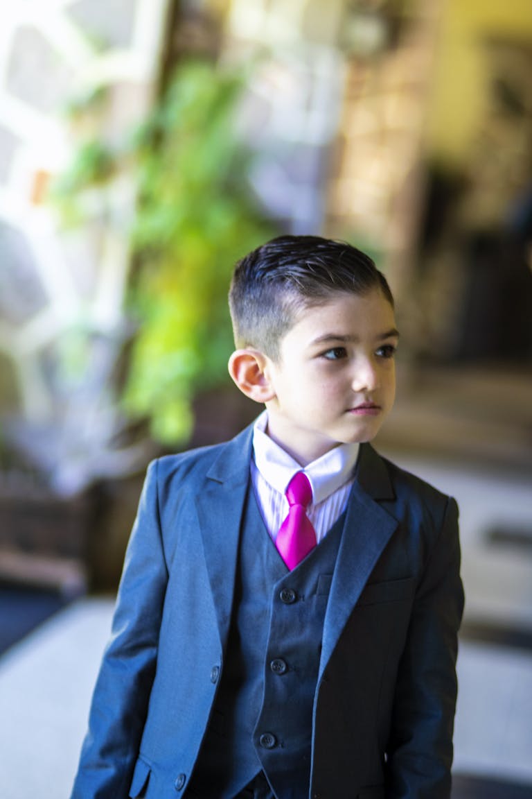 Young boy in a formal suit with a pink tie indoors, looking thoughtful.