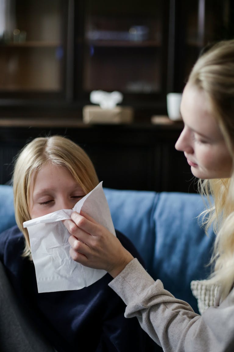 A mother gently wipes her child's nose with a tissue as they rest on a couch indoors.