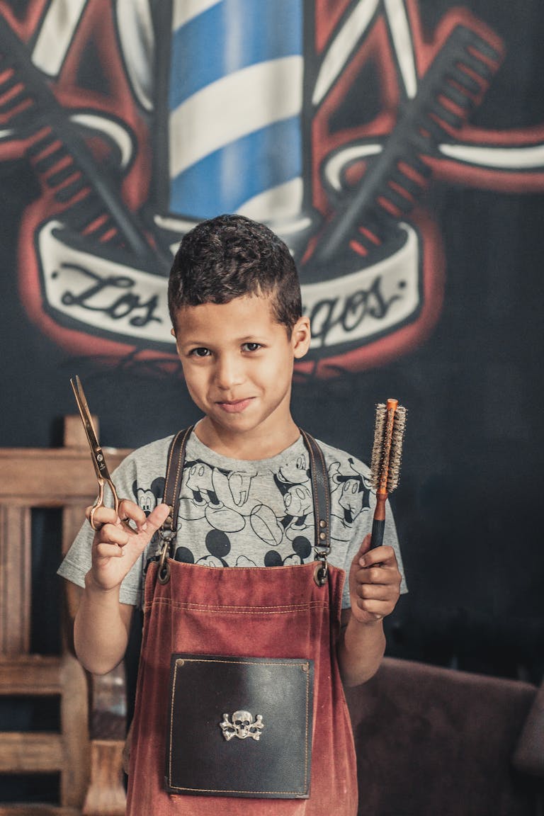Charming young boy dressed as a barber, holding scissors and a brush, indoors.