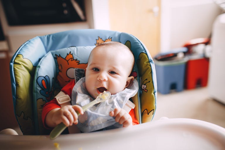 Cute little baby with mouth opened putting spoon with food in mouth while sitting in feeding chair