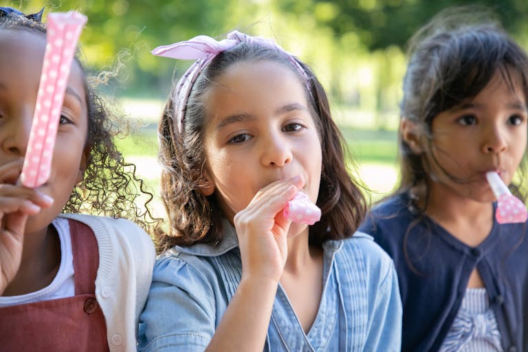 Three happy girls blowing party whistles outdoors, celebrating together in a park.
