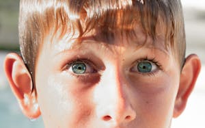 Intense close-up portrait of a young boy with striking blue eyes and wet hair, captured outdoors in bright sunlight.