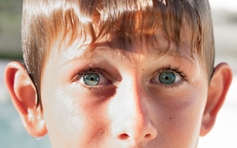 Intense close-up portrait of a young boy with striking blue eyes and wet hair, captured outdoors in bright sunlight.